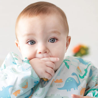 A baby in Bumkins Ultimate Gift Set - Dinosaurs & Blue Tropic, finger in mouth, eagerly readies for self-feeding adventures.