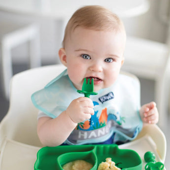 Baby in a high chair, wearing a bib, using Bumkins Ultimate Gift Set - Dinosaurs & Blue Tropic to eat independently.