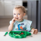 A curly-haired toddler self-feeds at the table using Bumkins Ultimate Gift Set - Dinosaurs & Blue Tropic, featuring a dinosaur bib and placemat.