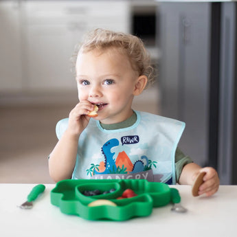 A curly-haired toddler self-feeds at the table using Bumkins Ultimate Gift Set - Dinosaurs & Blue Tropic, featuring a dinosaur bib and placemat.