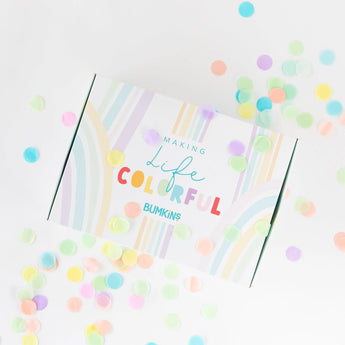 Surrounded by pastel confetti, the Bumkins Ultimate Gift Set - Ocean Life & Whale Tail features a Silicone First Feeder. The package is a white box adorned with colorful pastel stripes and the words Making Life Colorful Bumkins, adding charm to any meal.