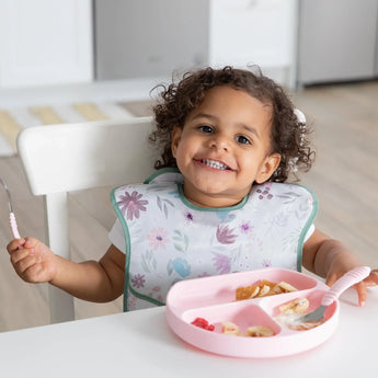 A smiling toddler, wearing a Bumkins Ultimate Gift Set - Floral & Lace, holds Chewtensils and eats from a pink Silicone Grip Dish.