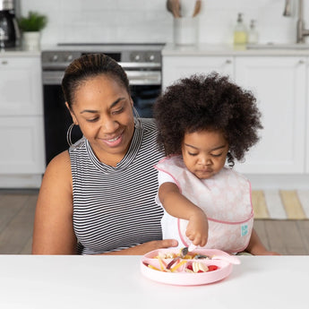 A woman smiles at a toddler using the Ultimate Gift Set - Floral & Lace from Bumkins, eating from a pink Silicone Grip Dish.