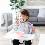 A toddler delights in a meal with the Bumkins Ultimate Gift Set - Floral & Lace. A sofa and plant are blurred in the background.