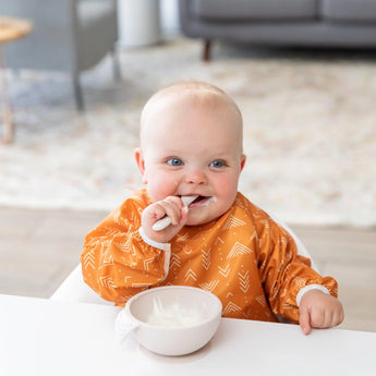 Baby in orange SuperBib from Bumkins Ultimate Gift Bundle, Desert Boho self-feeds with a spoon at the table using a Silicone Grip Dish.