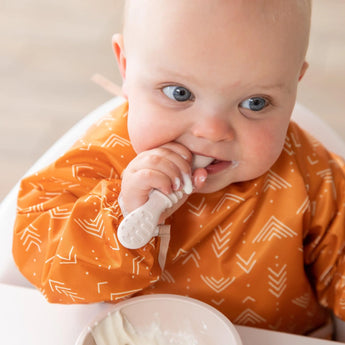 Baby in Bumkins Ultimate Gift Bundle, Desert Boho, chews on spoon beside a Silicone Grip Dish with yogurt for self-feeding fun.