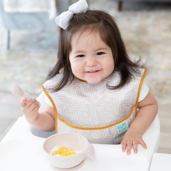 A toddler with a bow and SuperBib from the Bumkins Ultimate Gift Bundle smiles, spoon in hand, ready to eat from a Silicone Grip Dish.