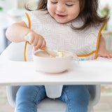 A toddler in a Bumkins Desert Boho Ultimate Gift Bundle self-feeds with a spoon from a Silicone Grip Dish.