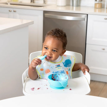 A smiling baby self-feeds with Bumkins Ultimate Gift Set in a cozy kitchen, wearing colorful gear from the Ocean Life & Whale Tail series.