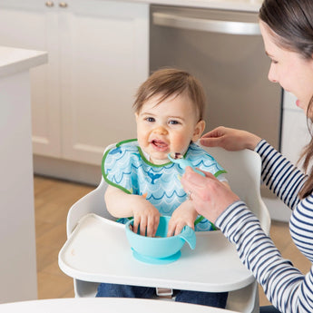 A baby in a high chair, sporting Bumkins Ultimate Gift Set - Ocean Life & Whale Tail, smiles with food around its mouth. The baby plays with Chewtensils while a woman beside them smiles, holding a spoon to feed or interact. A cozy kitchen completes the backdrop.