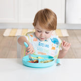 A toddler with light brown hair uses Bumkins Ultimate Gift Set to self-feed from a blue plate while wearing a SuperBib at a white table.