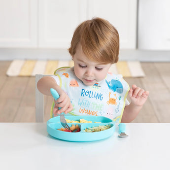 A toddler with light brown hair uses Bumkins Ultimate Gift Set to self-feed from a blue plate while wearing a SuperBib at a white table.