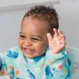 Baby smiling, hands raised, wearing a colorful Bumkins SuperBib from the Ultimate Gift Set - Ocean Life & Whale Tail, ready to self-feed!.