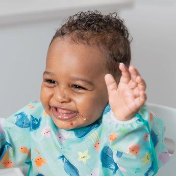 Baby smiling, hands raised, wearing a colorful Bumkins SuperBib from the Ultimate Gift Set - Ocean Life & Whale Tail, ready to self-feed!.