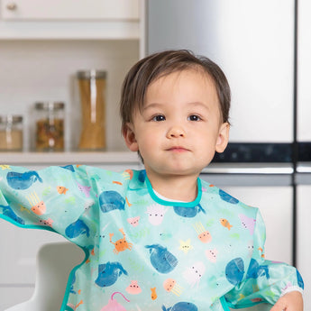 A toddler in a colorful Bumkins SuperBib from the Ultimate Gift Set - Ocean Life & Whale Tail sits attempting self-feeding.