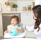 A smiling toddler in a high chair uses Bumkins Ultimate Gift Set as a woman with long dark hair sits beside them in the kitchen.