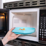 A hand places a blue Bumkins Silicone Grip Plate with food in a microwave.