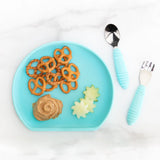 The Bumkins Silicone Grip Plate in Blue holds pretzels, peanut butter, and star apple; childs fork and spoon rest on a marble surface.