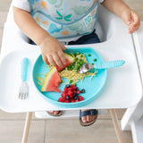 A child eats peas, watermelon, and pasta from a Bumkins Silicone Grip Plate: Blue. Toddler wears a dinosaur-print bib.