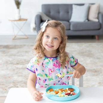 Curly-haired young girl in a floral shirt smiles while enjoying her meal from a blue Bumkins Silicone Grip Plate at the table.