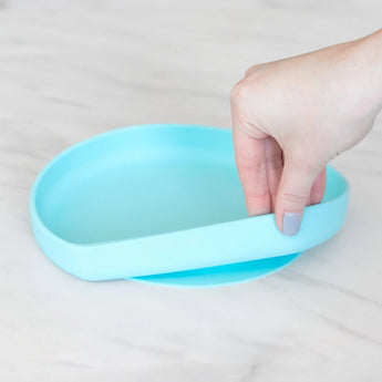 A hand with painted nails grips the edge of a Bumkins Silicone Grip Plate: Blue on a marble surface.