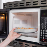 A hand places a Bumkins Silicone Grip Plate: Gray, crafted from food-safe silicone, inside the microwave.