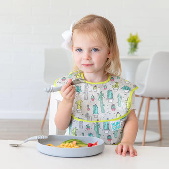 A toddler with a bow happily eats macaroni and strawberries from a food-safe Bumkins Silicone Grip Plate, Gray, wearing a cactus bib.