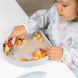 A toddler in a floral bib enjoys pasta and veggies from a Bumkins Silicone Grip Plate: Gray, its suction base keeps everything secure.