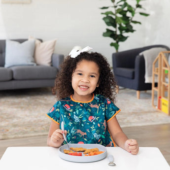 A smiling girl with curly hair enjoys chicken nuggets from a Bumkins Silicone Grip Plate: Gray at a white table in her cozy living room.