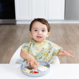 A toddler with a bib joyfully grips a bagel over a Bumkins Silicone Grip Plate: Gray, surrounded by various snacks.