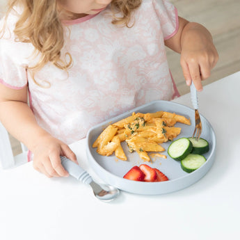 A child enjoys pasta, cucumber, and strawberries from a gray Bumkins Silicone Grip Plate with a secure suction base.