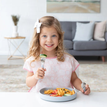 A curly-haired girl smiles, holding a fork over her Bumkins Silicone Grip Plate: Gray filled with pasta.