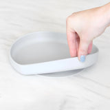 A hand lifts a gray Silicone Grip Plate by Bumkins from the light marble surface.