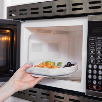 A person places a colorful pasta dish with broccoli and olives into the microwave using the Bumkins Silicone Grip Plate: Marble.