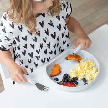 A child enjoys scrambled eggs, berries, and sweet potatoes from a Bumkins Silicone Grip Plate: Marble while wearing a shirt with black hearts.