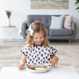 A girl enjoys fruit and cheese on a Bumkins Silicone Grip Plate: Marble at a white table in a cozy living room.