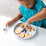 A child in a teal shirt enjoys snacks on a food-safe Silicone Grip Plate: Marble by Bumkins.