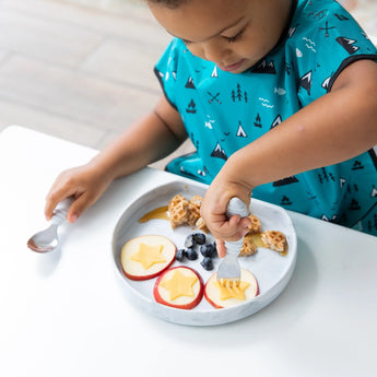 A child in a teal shirt enjoys snacks on a food-safe Silicone Grip Plate: Marble by Bumkins.