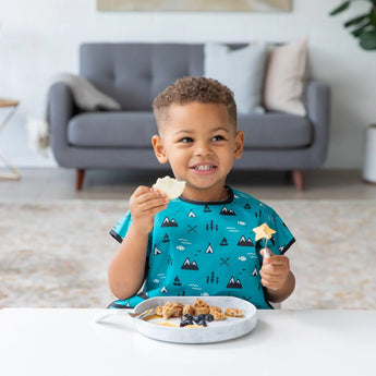 A child in a blue shirt smiles while eating from a Bumkins Silicone Grip Plate: Marble at the table with a sofa in the background.