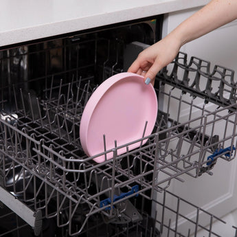 Someone puts a Bumkins Silicone Grip Plate: Pink, featuring a suction base, into a dishwasher rack.