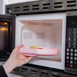 Placing a pink Bumkins Silicone Grip Plate of pasta into an open microwave.