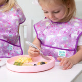 Two toddlers in purple bibs sit at a table. One uses a fork with a Bumkins Silicone Grip Plate in pink to eat scrambled eggs.