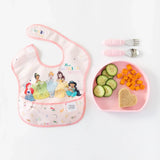 Bumkins Silicone Grip Plate: Pink with cucumbers, carrots & heart sandwich, next to a bib with cartoon princesses.