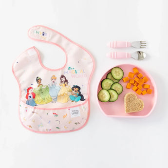 Bumkins Silicone Grip Plate: Pink with cucumbers, carrots & heart sandwich, next to a bib with cartoon princesses.