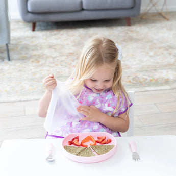 Young girl in a purple bib unwraps a sandwich on a Bumkins Silicone Grip Plate: Pink with strawberries, secured by its suction base.