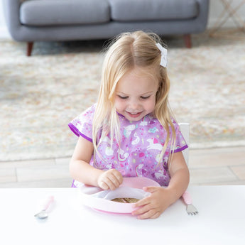 A girl in a purple bib smiles at the table holding a Bumkins Silicone Grip Plate: Pink. A spoon and fork rest nearby.