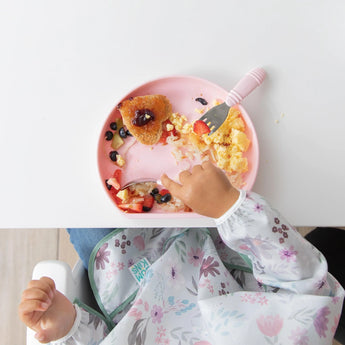 A child enjoys fruit and a nugget from a Bumkins Silicone Grip Plate: Pink, with a suction base, wearing a floral bib at a white table.