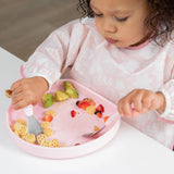 A toddler enjoys pasta, avocado, and strawberries from a Bumkins Silicone Grip Plate: Pink at the white table.