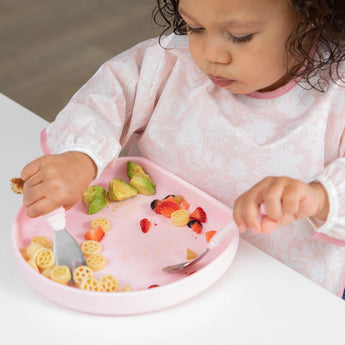 A toddler enjoys pasta, avocado, and strawberries from a Bumkins Silicone Grip Plate: Pink at the white table.