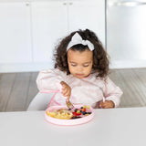 A toddler enjoys scrambled eggs and berries on a pink silicone grip plate by Bumkins, sitting in a white kitchen.